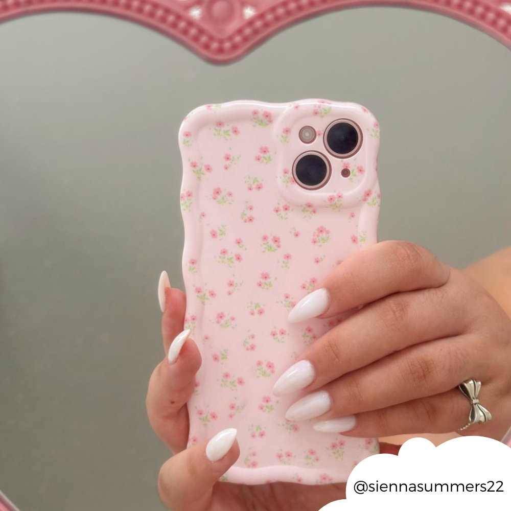 Hand with long white nails holds a pink, wavy floral phone case in front of a heart-shaped mirror.
