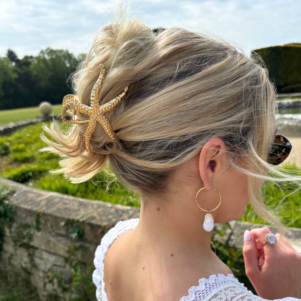 Blonde hair in an updo with a gold metal starfish claw clip, paired with gold hoops and pearl drop earrings.