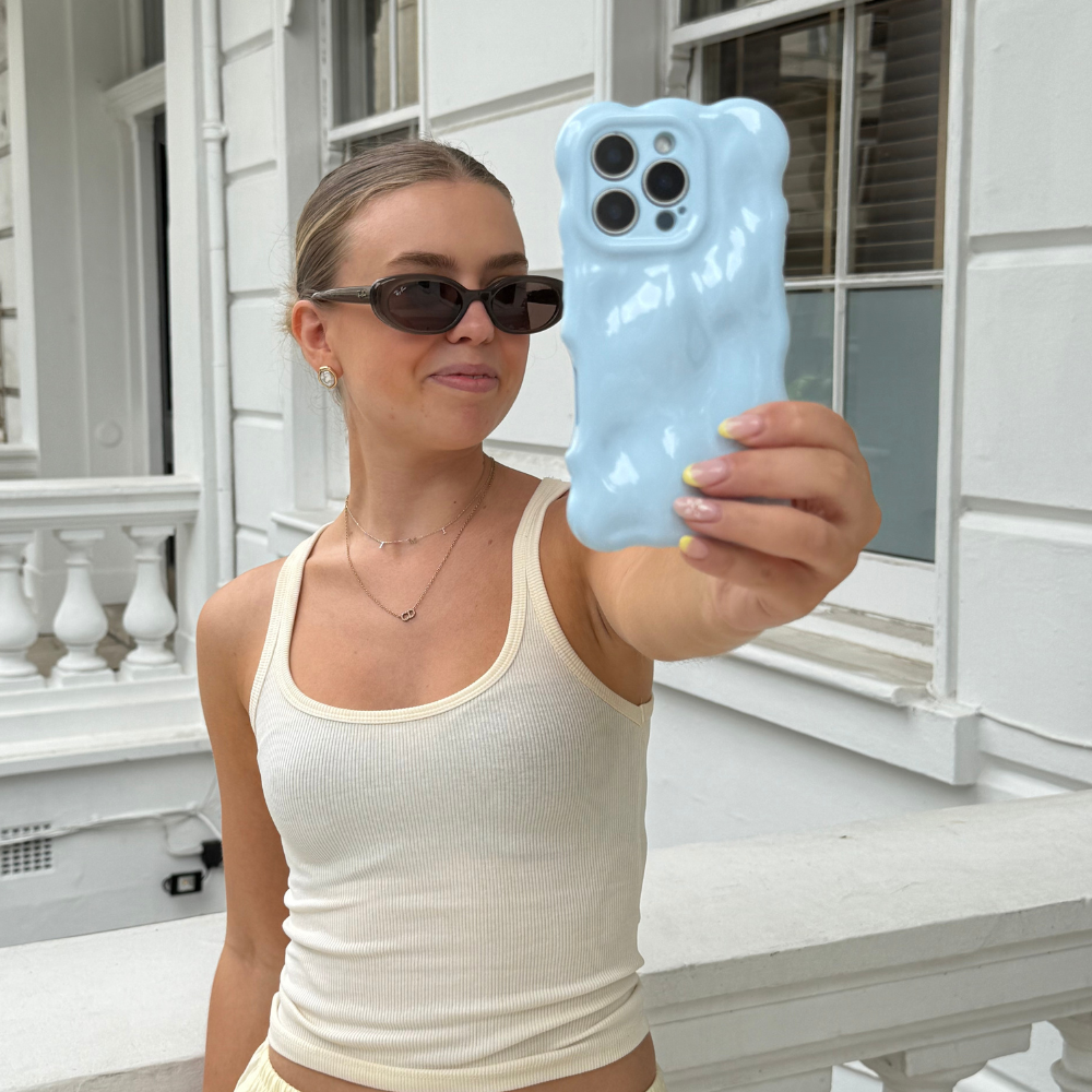 Woman in a cream tank top holding a baby blue 3D bubble phone case by Coconut Lane near a white building.