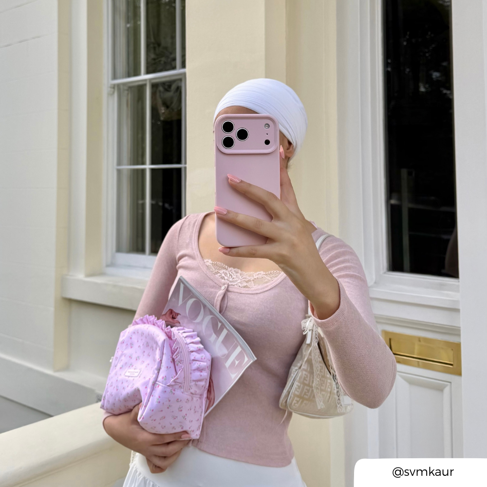 A woman takes a selfie outdoors in front of a cream-colored building. She is wearing a light pink long-sleeve top, a white headwrap, and white bottoms, holding an iphone with a light pink phone case. She is holding a ditsy floral pink makeup bag and vogue magazine.