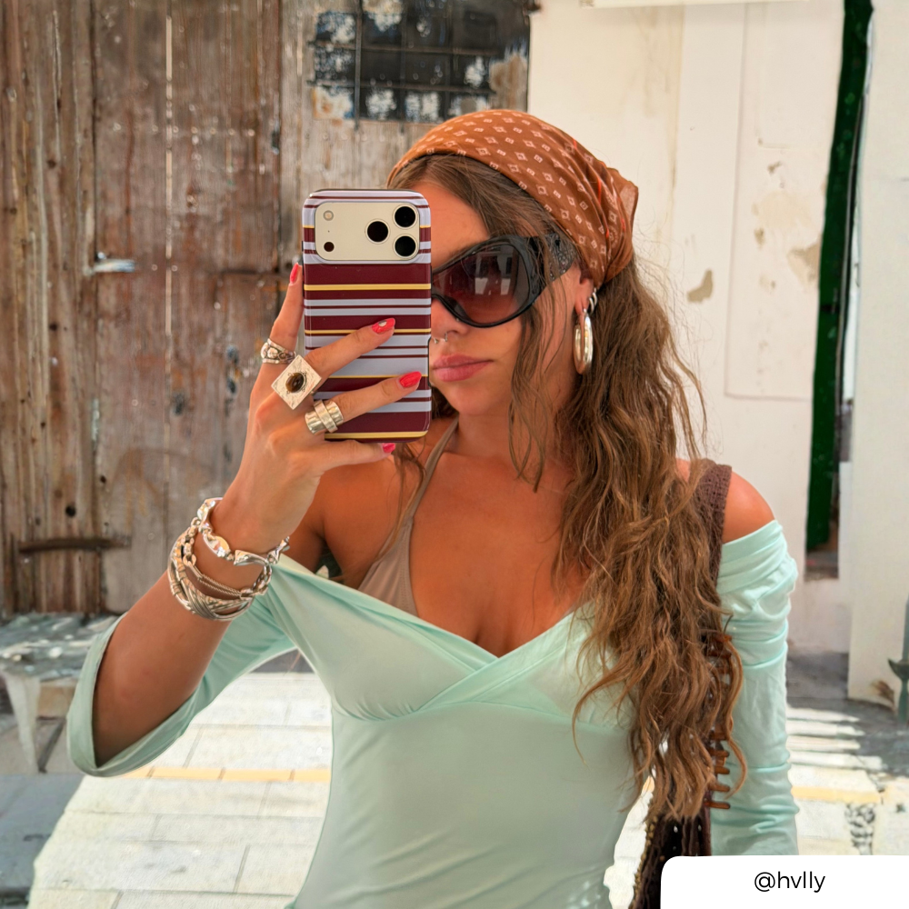 A woman takes a mirror selfie wearing a light mint green off-the-shoulder long-sleeve top, a brown patterned headscarf, dark sunglasses, and multiple rings and bracelets. She is standing outdoors in front of a rustic wooden door and white wall. She is holding an iphone with a striped brown, blue and yellow phone case.