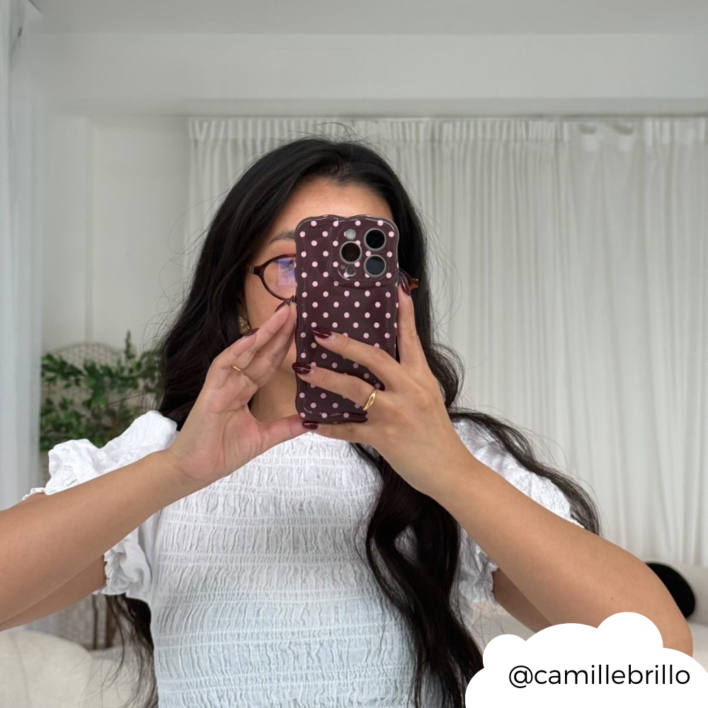 Woman in a white top holds up a brown polka dot wavy phone case, partly covering her face in a mirror selfie.