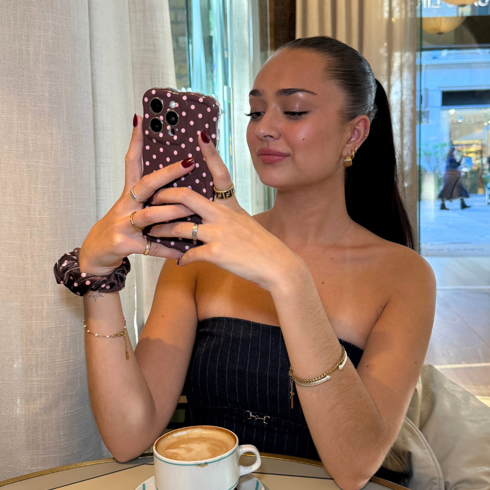 Woman with ponytail at café table smiles at her phone in a brown polka dot Coconut Lane wavy phone case.
