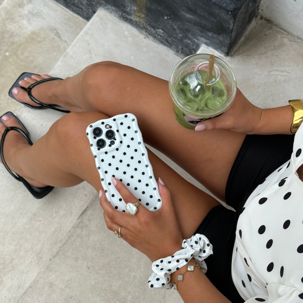 Woman in coordinated outfit holds a green drink and phone with white polka dot wavy case from Coconut Lane.