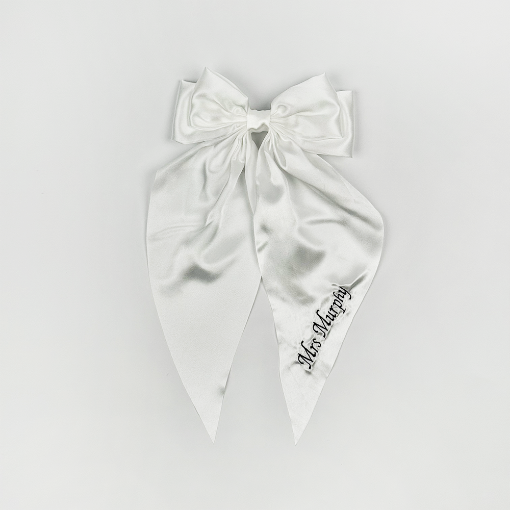 A large white satin bow with long tails, featuring black embroidered text that reads "Mrs. Murphy" on one of the tails, displayed against a plain white background.