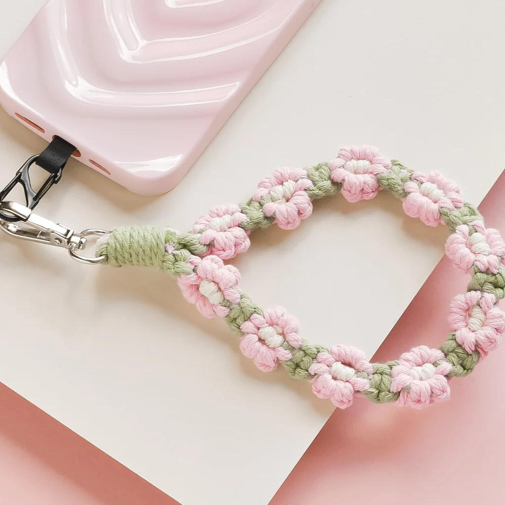 A pastel pink and green floral handwoven phone strap attached to a pink phone case, placed on a light pink and white background. The strap features small flowers interwoven in a braided pattern.