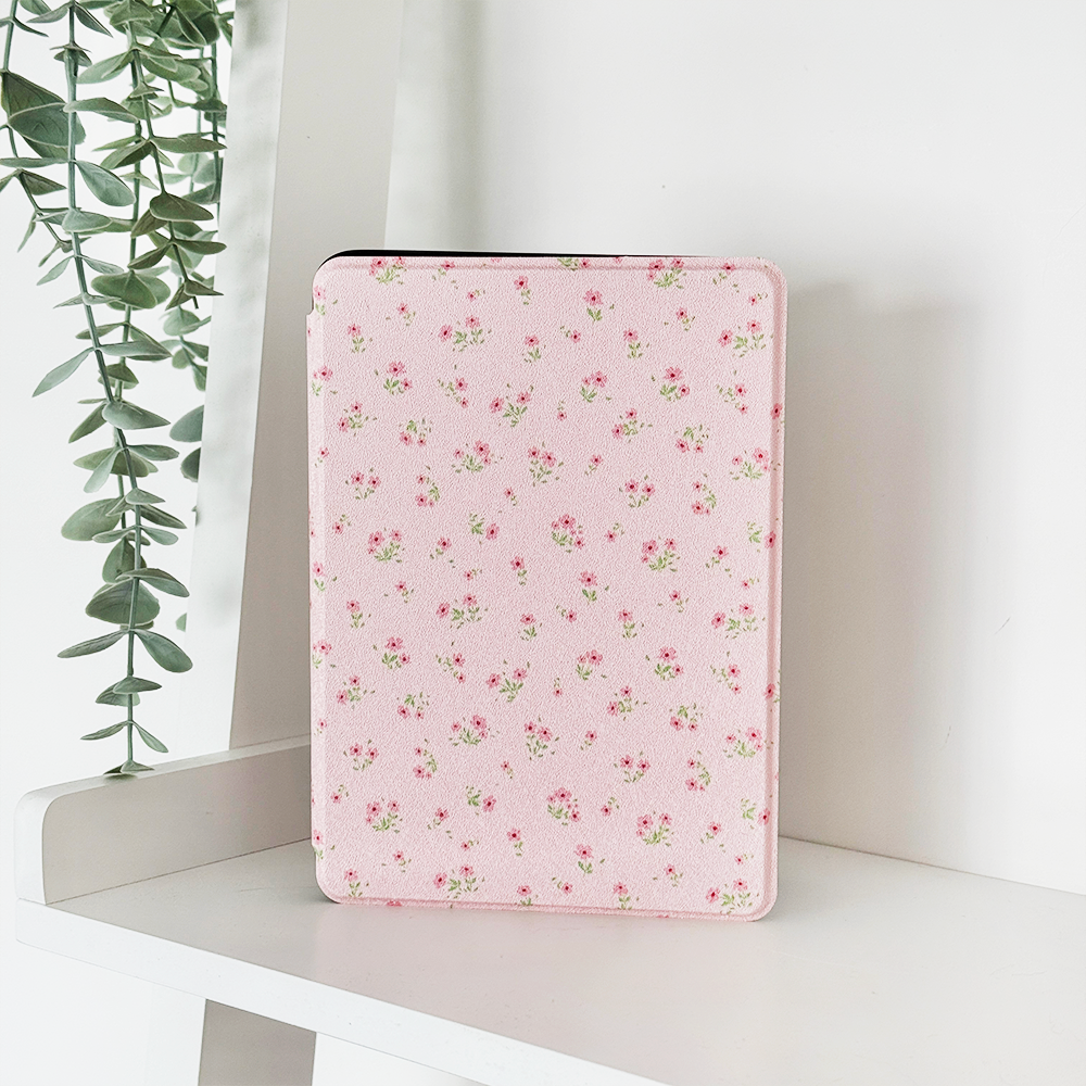 A white tablet case with a pink bow pattern stands upright on a white shelf, with a green trailing plant in the background.