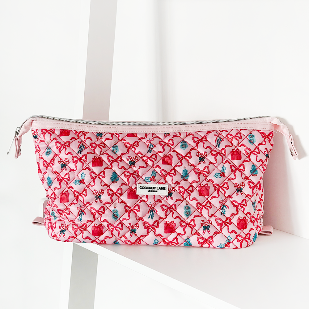 A quilted pink pouch with a pattern of red hearts and small flowers, featuring a white zipper and a "Coconut Lane" label at the front, placed on a white surface.