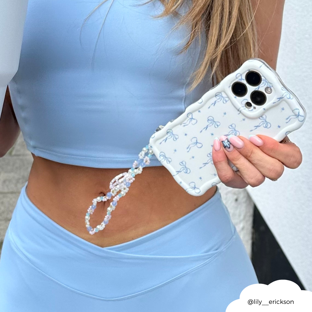 Woman in light blue outfit holds iPhone with Coconut Lane beaded phone strap and Ditsy Flower Bow patterned case.