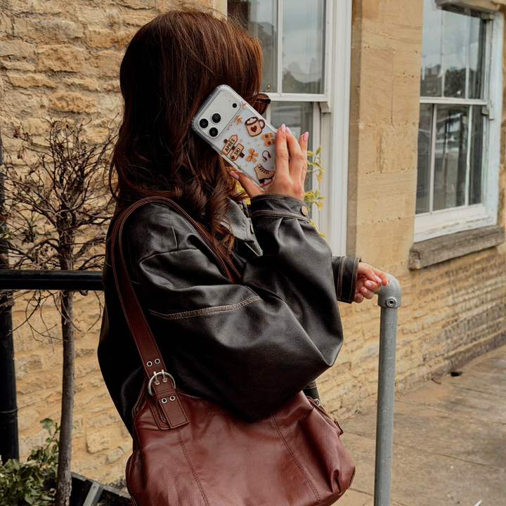 A woman in a dark brown leather jacket and brown leather shoulder bag stands outdoors by a stone building, holding a phone with a clear phone case with decorative winter illustrations including a coffee mug, a pretzel, ice skate and a sign saying Apres Ski. 