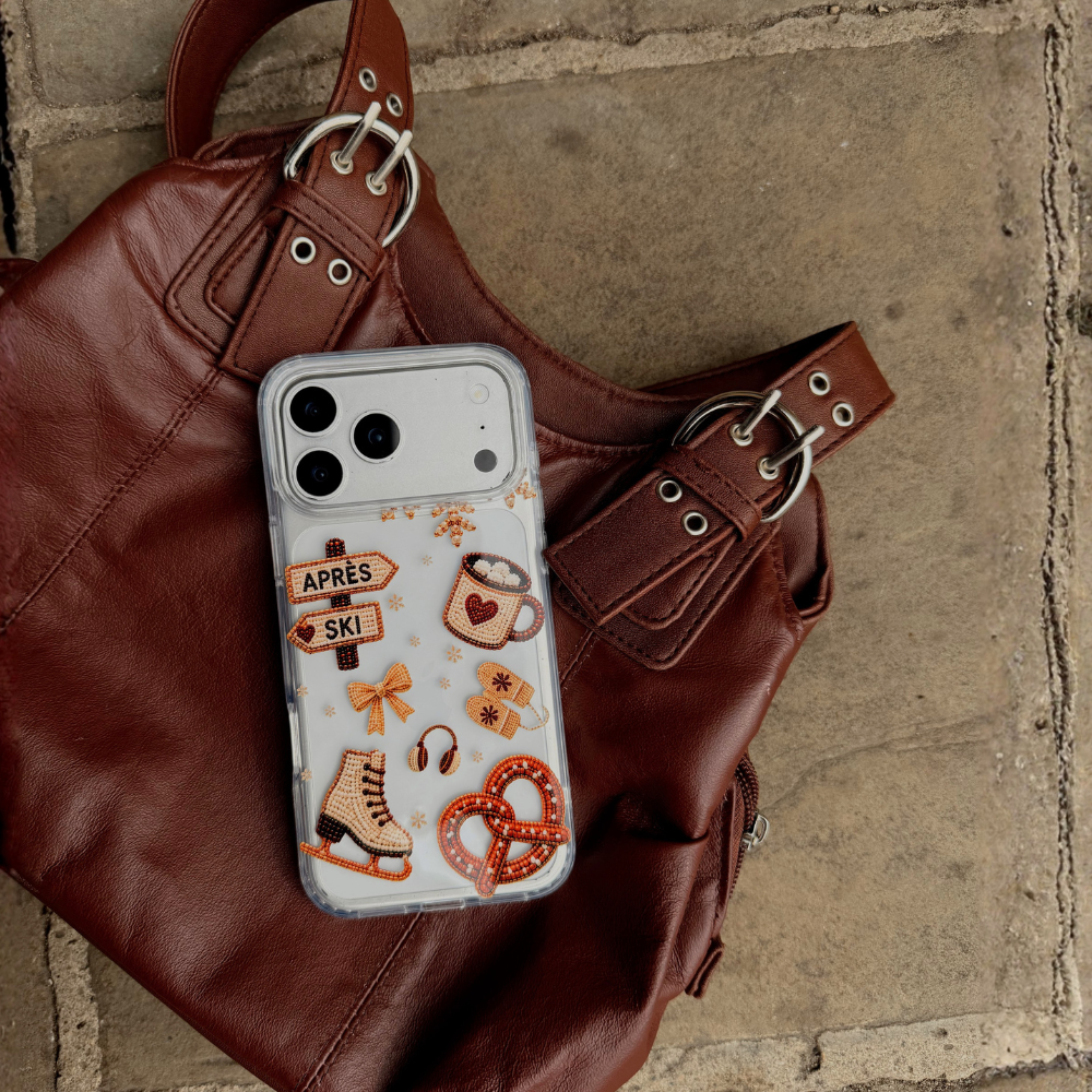 A clear phone case with winter and ski-themed illustrations, including a mug with a heart, a ice skate, and directional signs reading 'APRÈS' and 'SKI', shown on an iphone and placed on a brown leather handbag over a stone surface.