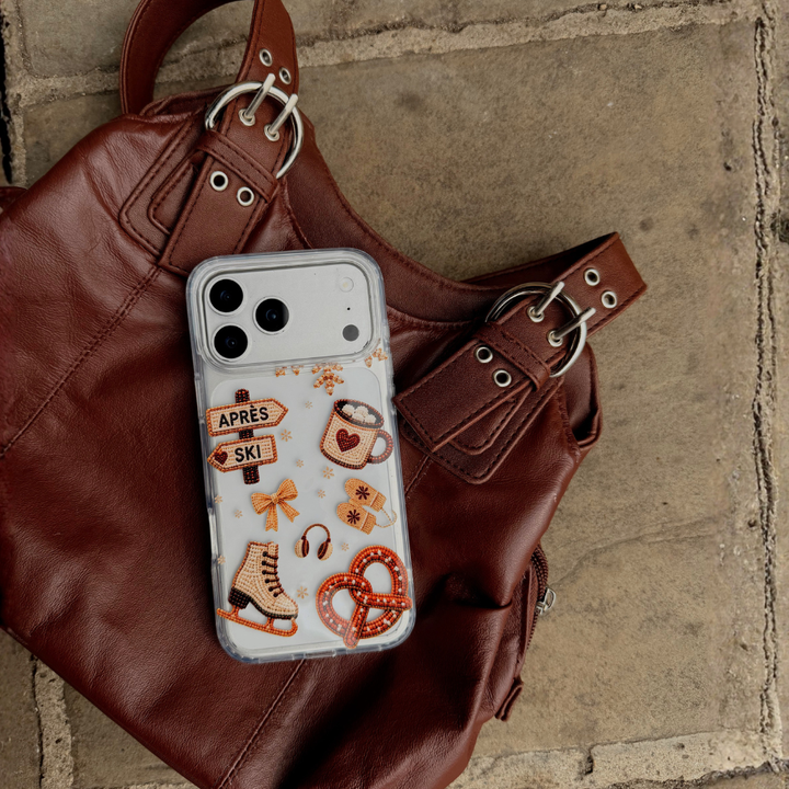 A clear phone case with winter and ski-themed illustrations, including a mug with a heart, a ice skate, and directional signs reading 'APRÈS' and 'SKI', shown on an iphone and placed on a brown leather handbag over a stone surface.