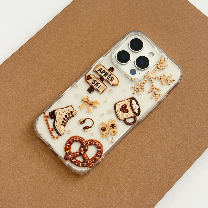 A clear phone case decorated with beige and brown ski-themed graphics, including skis, a pretzel, a coffee cup, a bow, and directional signs reading 'APRÈS' and 'SKI'.