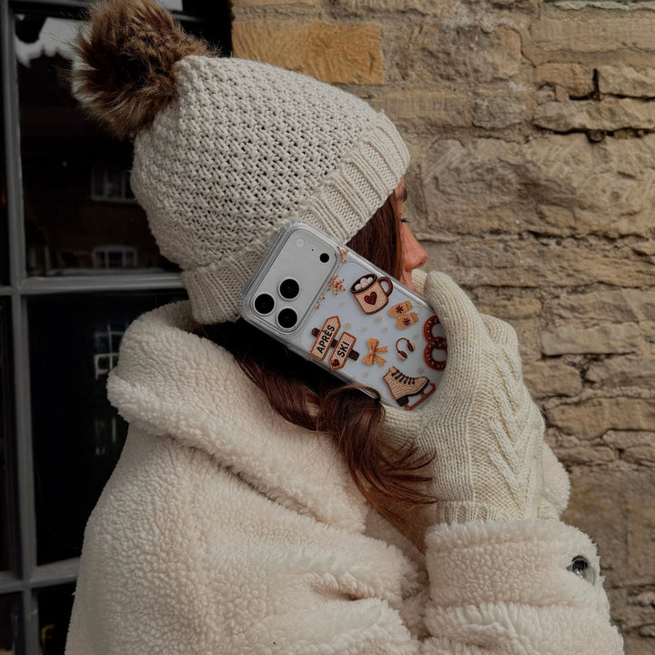 A woman wearing a cream knit beanie with a brown pom-pom and matching knit gloves holds a phone with a clear Apres Hour phone case.