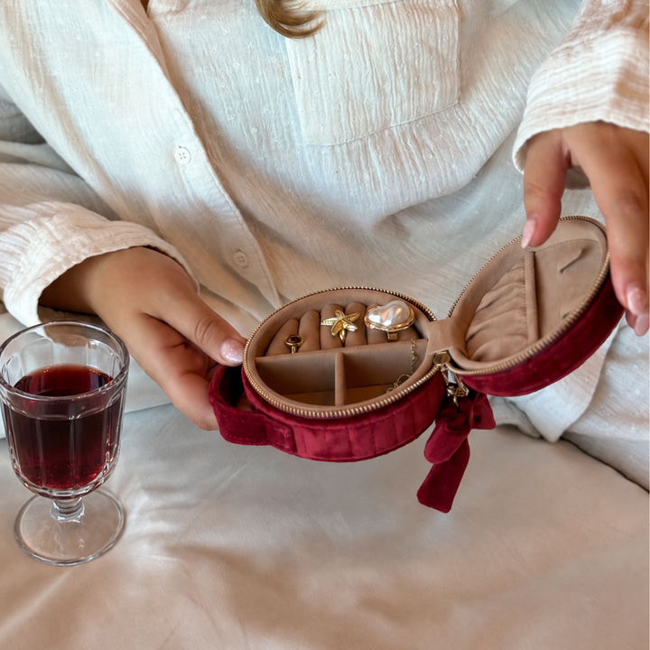 Burgundy velvet jewellery box with a quilted design, top handle, bow detail, and gold-tone hardware. Open with jewellery inside, next to a glass of red wine
