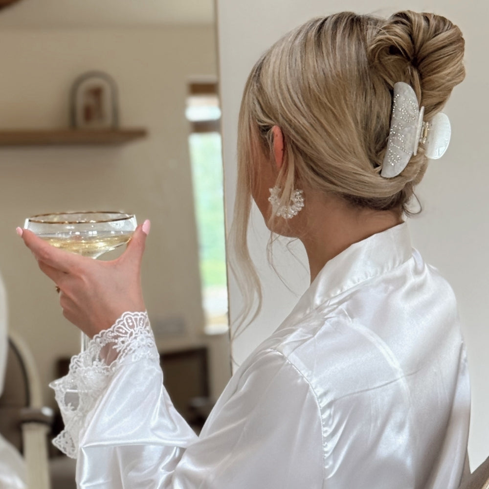 Girl in a white satin robe holding a glass of prosecco with bride claw clip
