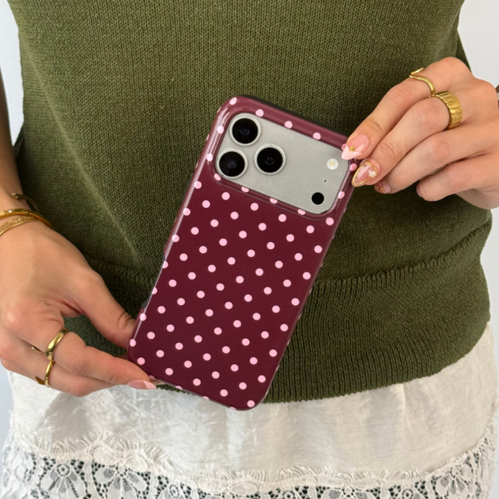 Woman wearing a dark green jumper with a white lace shirt underneath holds an iphone in a browny burgundy case with baby pink polka dots. 