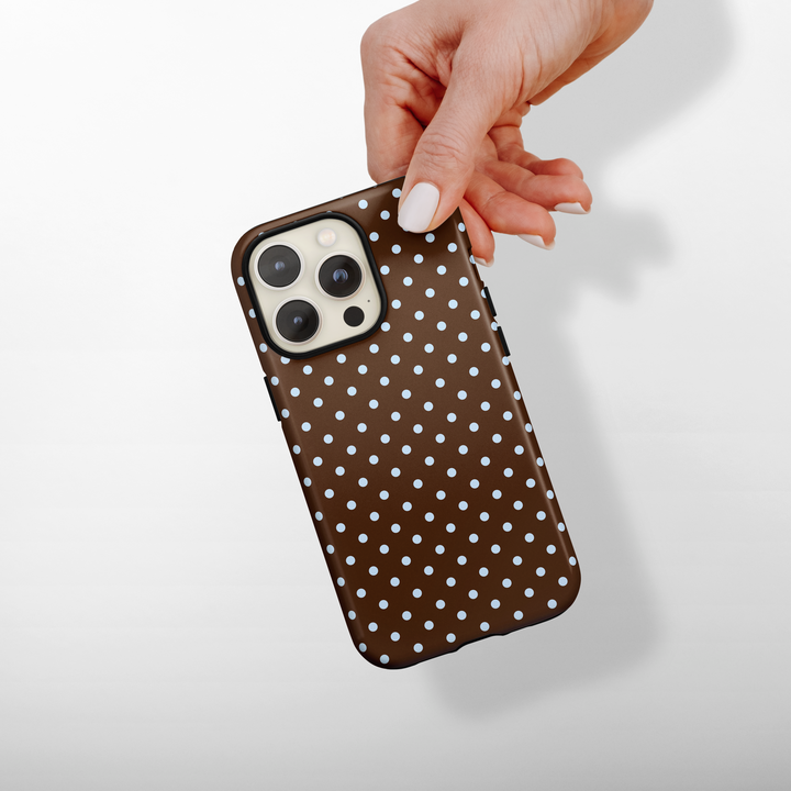 Hand holding a phone with a brown polka dot case on a light background