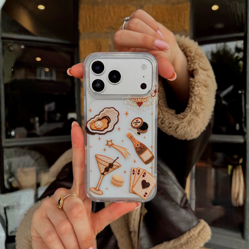 A person holds up a clear phone case decorated with gold-toned cocktail, playing cards, and clam with pearl illustrations.