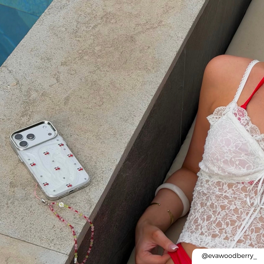 Phone case with red and white pattern cherries on a stone surface next to a person in a white lace dress.