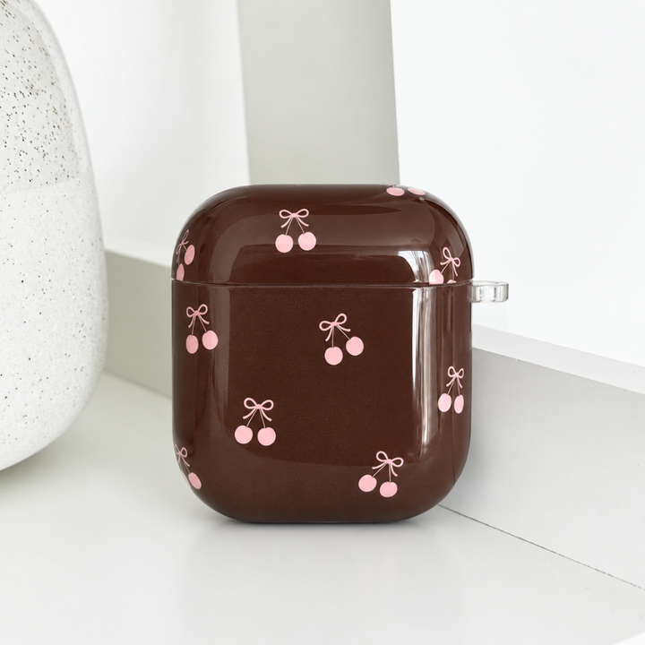 Glossy brown AirPods case with a pink cherry and bow print, closed and resting on a white shelf.