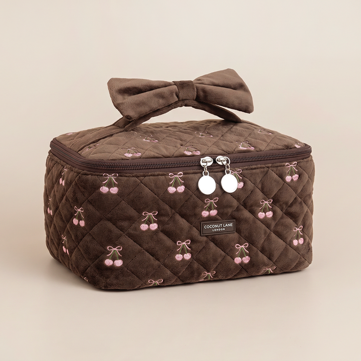 Brown quilted velvet vanity case with pink embroidered cherries, a velvet bow handle and a “Coconut Lane London” label.