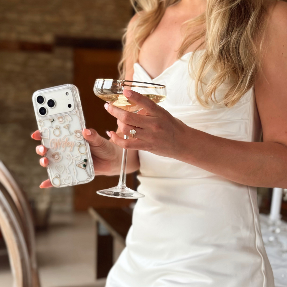 Woman in a white dress holding a clear wifey phone case and a glass of wine.