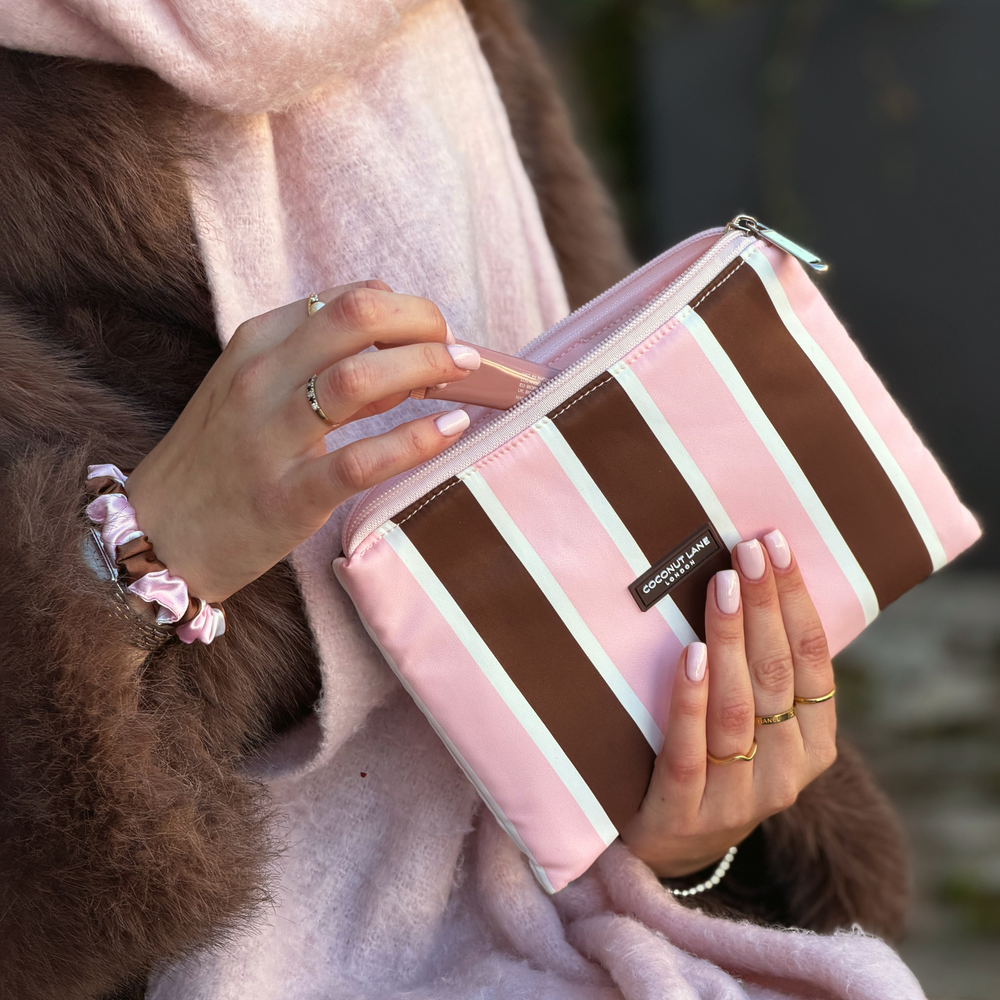 Woman holding the Coconut Lane Cocoa & Pink Stripe Everything Pouch featuring chocolate brown, pastel pink and white vertical stripes. On her wrist is the matching satin scrunchie. She is wearing a baby pink scarf and a faux fur brown jacket.