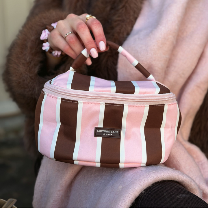 Woman with blonde hair, wearing a baby pink scarf and brown faux  fur jacket, holding a coconut Lane Cocoa & Pink Stripe round vanity makeup bag with chocolate brown and baby pink stripes.