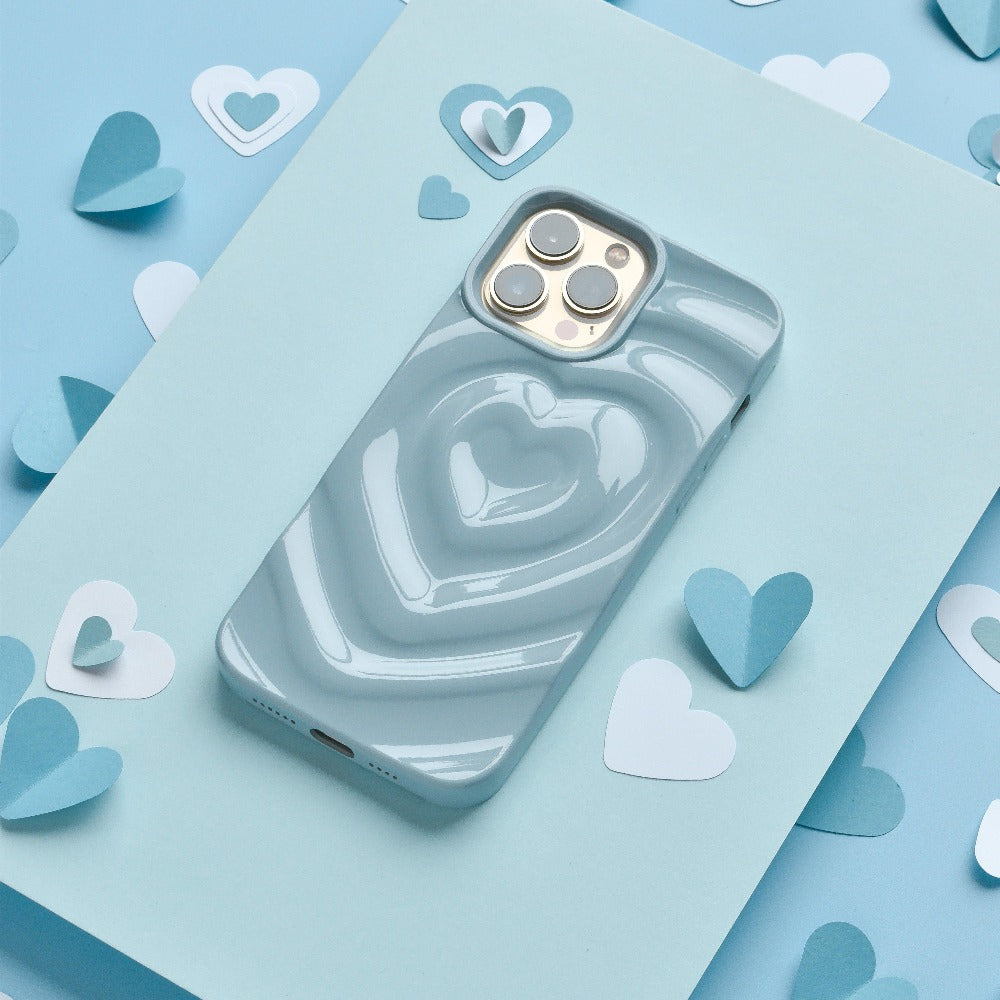 A smartphone adorned with Coconut Lane's "Melting Heart Phone Case - Baby Blue" lies on a light blue surface. Surrounding the phone are various paper cutouts of hearts in different shades of blue and white, enhancing the romantic theme of the image.