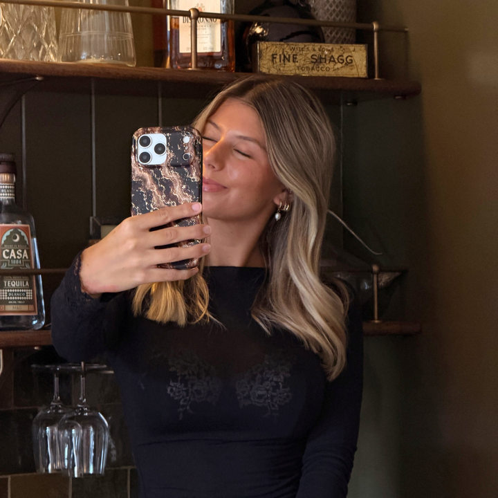 A woman takes a mirror selfie in a bar setting, wearing a black long-sleeve top, holding an  iphone in a beige and dark brown marble design with subtle gold glitter flecks phone case
