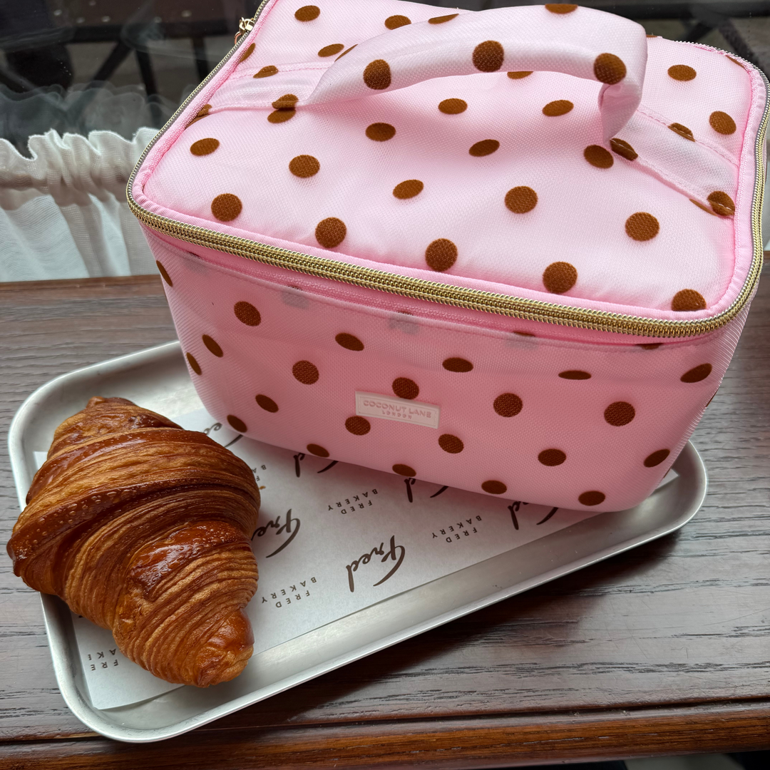 Flat lay photo in a cafe of the Dot Society pink and brown vanity case. The vanity case is baby pink mesh type fabric, with chocolate brown polka dots. 