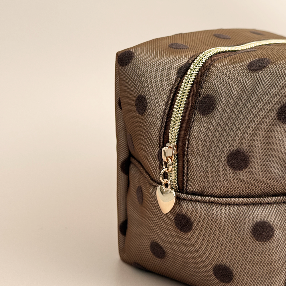 Close-up of brown mesh makeup bag with dark brown polka dots, a gold top zip with a heart-shaped zip pull, and a black Coconut Lane London logo badge on the front, shown on a beige background.