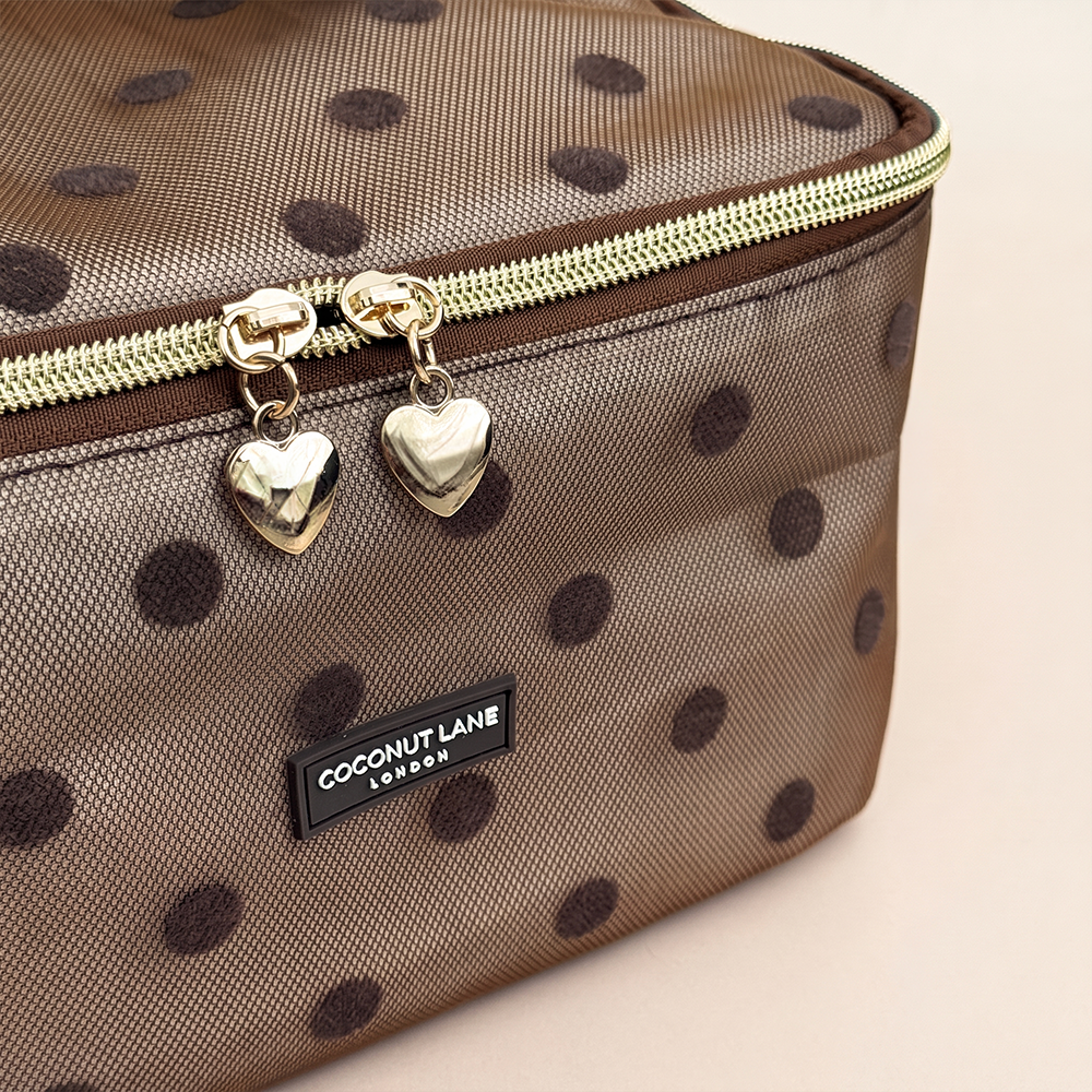 Close-up of brown mesh vanity case with dark brown polka dots, a padded top handle, gold zip with two heart-shaped zip pulls, and a black Coconut Lane London logo badge on the front, shown on a beige background.