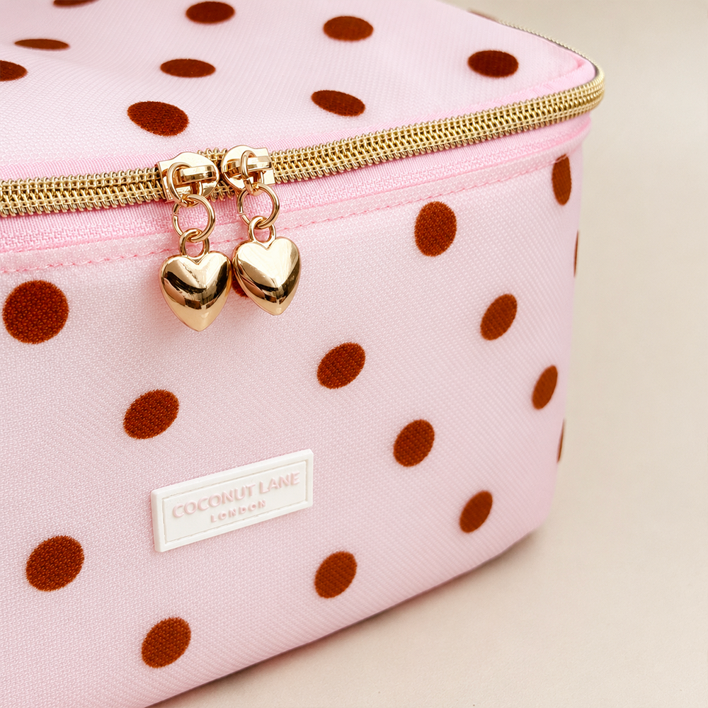 Close-up of pink mesh vanity case with brown polka dots, a padded polka dot top handle, gold zip with two heart-shaped zip pulls, and a white Coconut Lane London logo badge on the front, shown on a beige background.