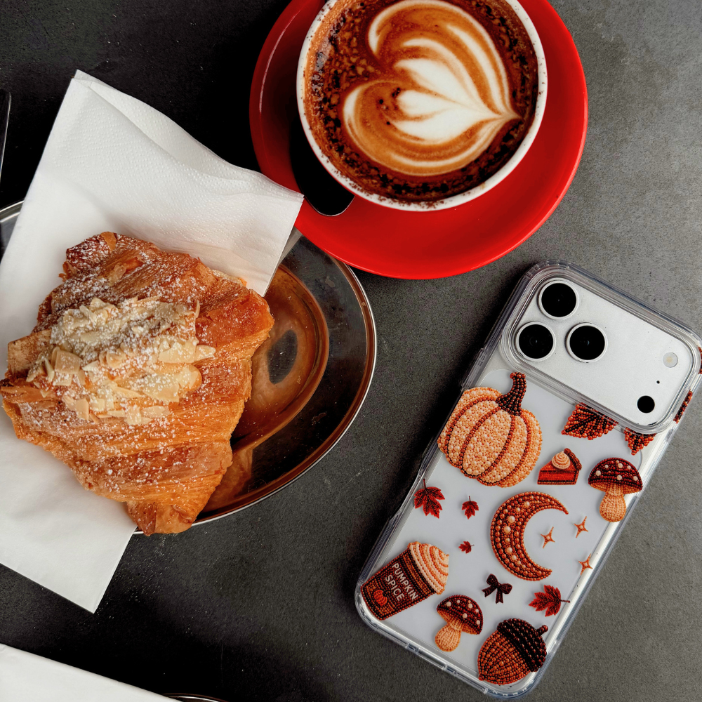 A clear phone case with an autumn-themed print of pumpkins, acorns, and mushrooms, alongside a croisant and a latte on a dark gray table.
