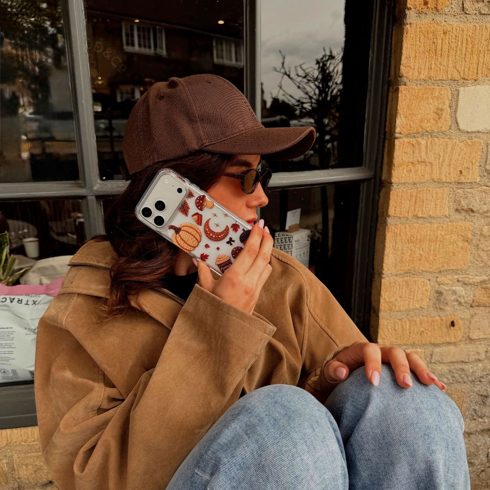 A woman wearing a brown cap, brown suede jacket, and blue jeans sits against a brick wall next to a window, holding a phone with autumn-themed phone case with pumpkins, acorns, mushrooms, leaves, and a pumpkin spice coffee cup, shown in a lifestyle cafe setting.
