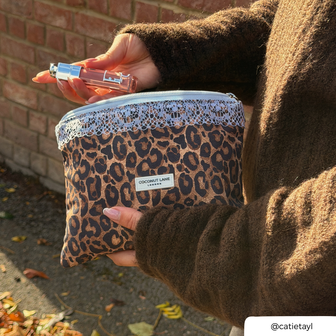 Woman in brown knit jumper, holding the leopard print zip pouch with a pale blue lace trim along the top, a pale blue zip with a silver pull, and a white Coconut Lane London logo badge on the front. The pouch is unzipped and she is pulling out a lipgloss. 
