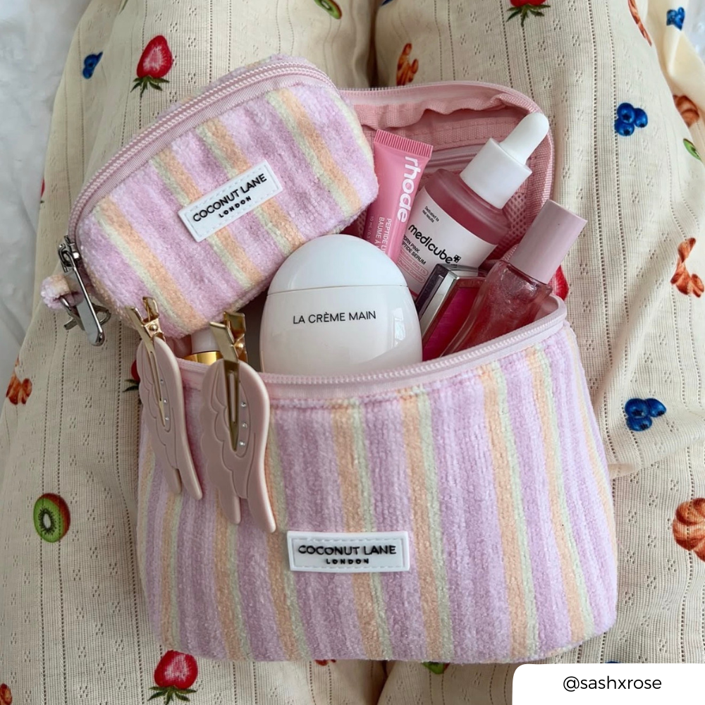 A pastel striped cosmetic pouch from Coconut Lane London sits open on a cream fabric surface with a strawberry and kiwi print. Inside are several skincare items, including a white La Creme Main container and pink Medipod bottles.