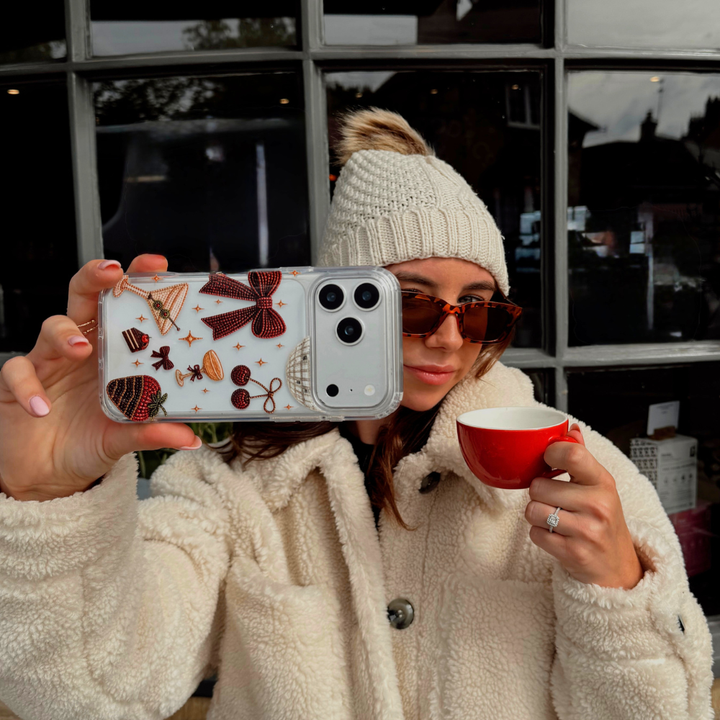 A woman wearing cream knit gloves and a cream teddy coat, holds up a clear phone case decorated with a red bow, cherries, a martini glass, a strawberry dipped in chocolate, and small star accents. The background shows a cafe setting.