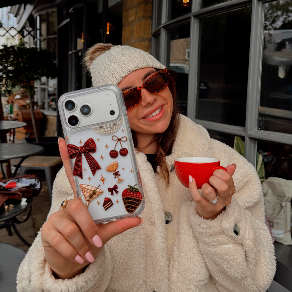 A woman wearing cream knit gloves and a cream teddy coat, drinking a coffee, holds up a clear phone case decorated with a red bow, cherries, a martini glass, a strawberry dipped in chocolate, and small star accents. The background shows a cafe setting.