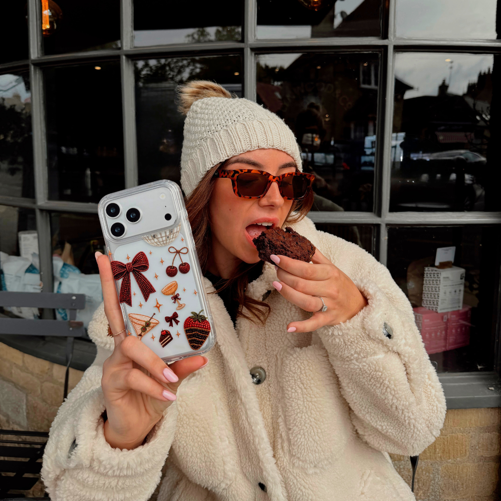 A woman wearing cream knit gloves and a cream teddy coat, taking a bite from a brownie, holds up a clear phone case decorated with a red bow, cherries, a martini glass, a strawberry dipped in chocolate, and small star accents. The background shows a cafe setting.