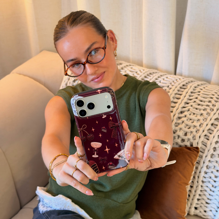 Woman in a dark green jumper with a white lace shirt underneath holds a burgundy chocolate brown phone case with decorative beaded illustrations on it, such as a bow, a strawberry, a cocktail, stars and cherries. 