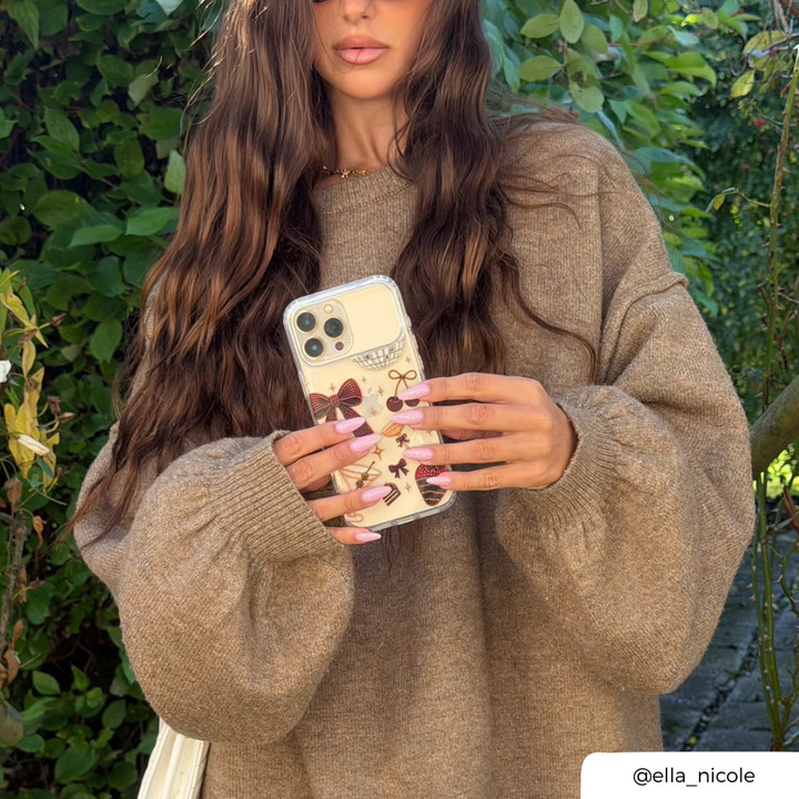 Woman in brown jumper, holding iphone with clear phone case decorated with a red bow, cherries, a martini glass, a strawberry dipped in chocolate, and small star accents. 