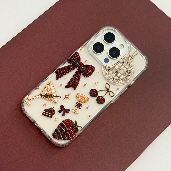 A clear phone case decorated with party-themed graphics including a large burgundy bow, a martini glass, cherries, a strawberry, a disco ball, and small gold stars.