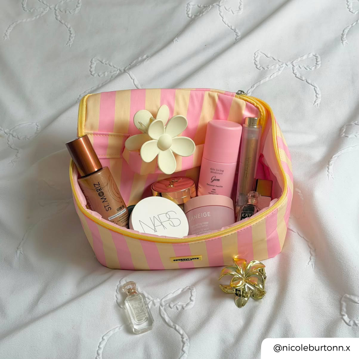 a flat lay photo of a pink and yellow vanity makeup bag with a butter yellow flower claw clip, and products showing inside the bag. The vanity case is placed on white bedding with white bow details.