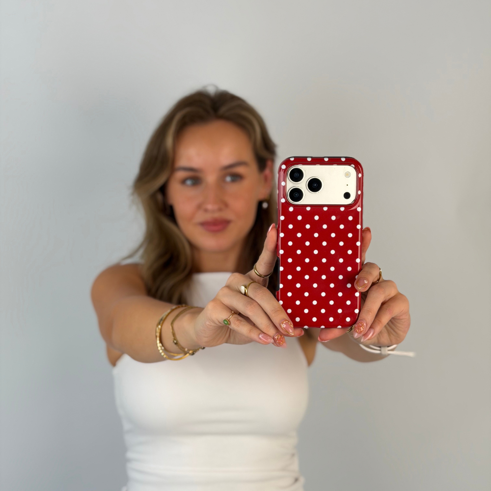Woman holding an iphone talking a selfie. The phone is in a red and white polka dot phone case. The woman is wearing a white vest top with gold jewellery. 
