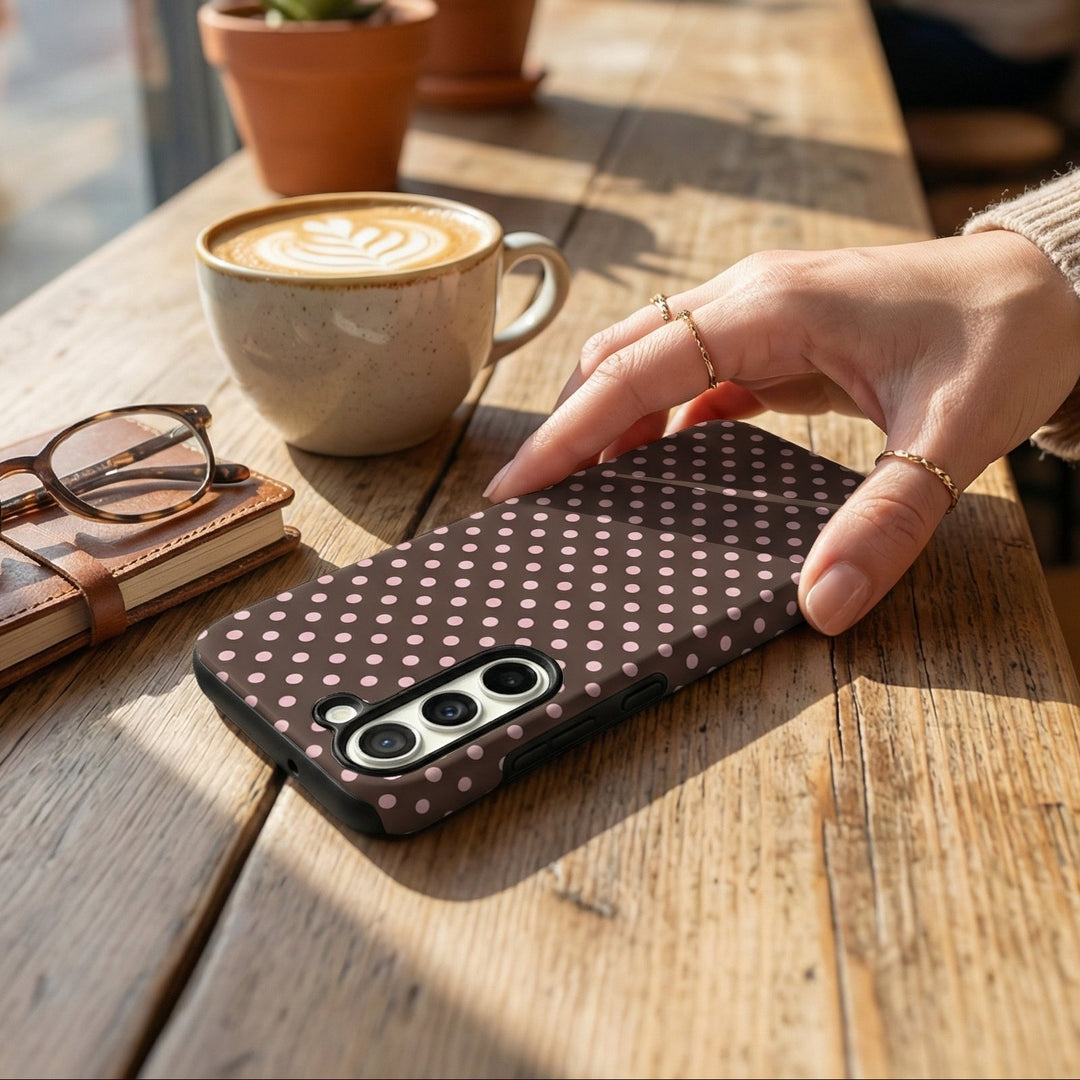 Person holding a phone with a polka dot case on a wooden table in a cafe.