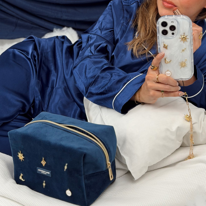 Woman wearing satin navy blue pyjamas holding a Stardust After Dark makeup bag. Dark navy blue velvet material with gold zip, and sewn-on gold celestial-themed charms including a sun, moon, star, diamond shape and 11:11 charm.  