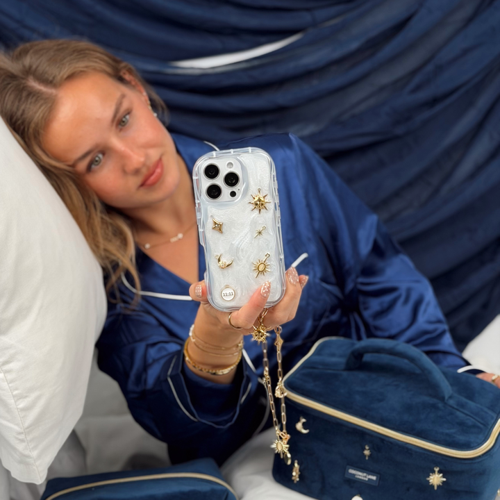 Woman wearing satin navy pyjamas laying in a bed with white bedding, She is holding a Stardust after dark charm phone case. A pearl base with luxe gold celestial charms including a moon, sun, star, diamond shape and 11:11 charm. Attached to the phone case is a matching gold chain phone strap. 